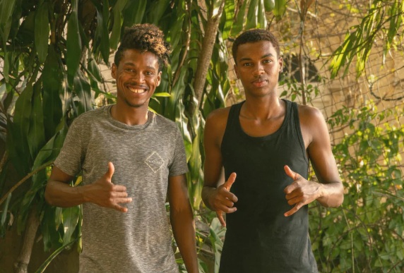 Two young men standing outdoors in a wooded area, smiling and giving a thumbs-up gesture to the camera.